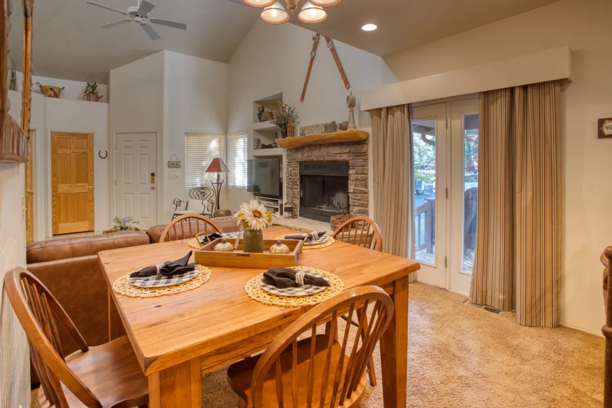 A large wooden dining table is positioned near floor-to-ceiling windows that flood the space with natural light. A stone fireplace anchors the room, while the open layout allows for comfortable family meals or lively group celebrations. The overall mood is bright and welcoming, perfect for both sunny brunches and snug winter nights.