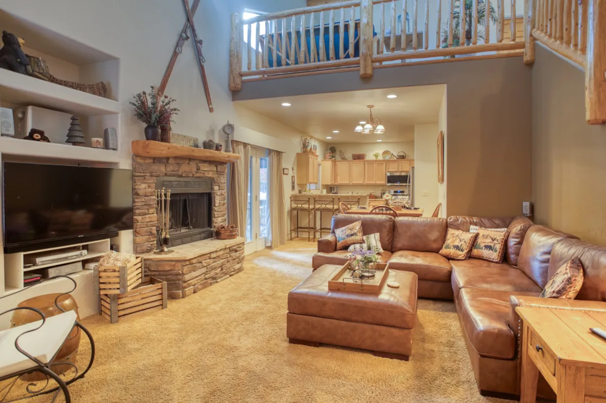 The image showcases a cozy, inviting great room with a striking stone fireplace as the centerpiece, a large leather sectional arranged for relaxed gatherings, and a modern kitchen visible in the background. The warm lighting and spacious layout create an atmosphere perfect for family time, entertaining, and unwinding after mountain adventures.