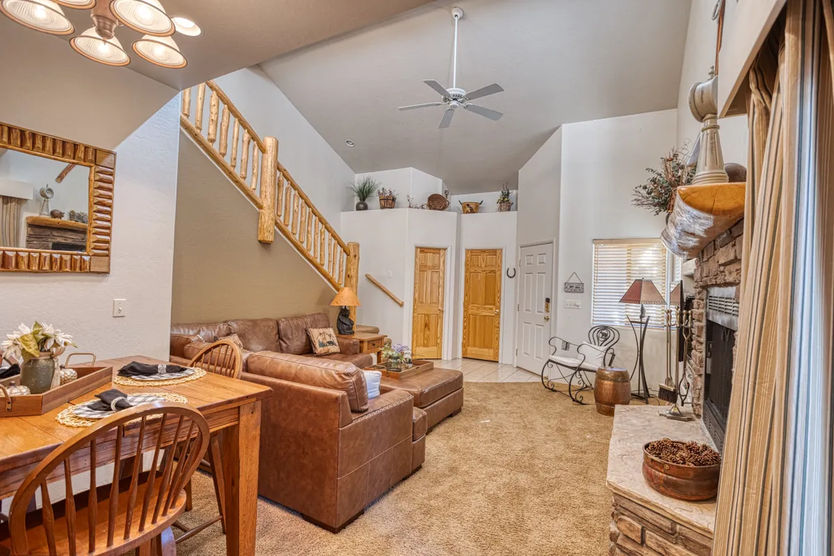 This image features a spacious great room with a leather sofa arranged for conversation, a wooden dining table set for gatherings, a prominent fireplace that serves as a cozy focal point, and a staircase that adds architectural interest. The warm color palette and soft lighting create a welcoming atmosphere ideal for family and friends to relax or entertain.